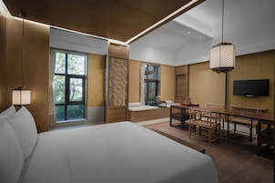 A modern hotel room with a large bed, a dining area with wooden furniture, a TV mounted on the wall, and large windows offering a view of the outdoors.