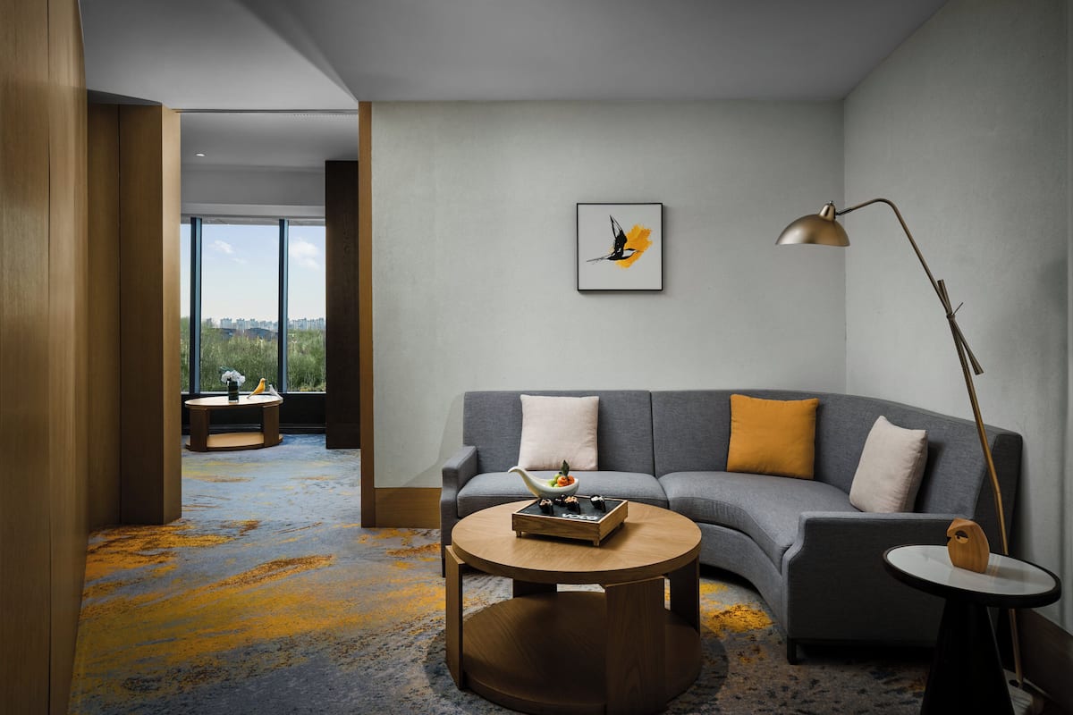 Una sala de estar moderna con un sofá gris, una mesa de centro redonda y una lámpara de pie. Hay una obra de arte enmarcada en la pared y una vista de la ciudad a través de ventanas amplias.
