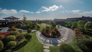 Un jardín tradicional coreano con un sendero circular central, rodeado de arbustos y árboles cuidadosamente podados, y arquitectura coreana tradicional al fondo.