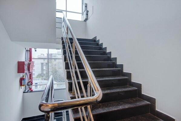A staircase with a metal handrail and black steps, leading to a landing with a window and a fire extinguisher mounted on the wall. - Botanica Luxury Living Space, Gachibowli, Hyderabad (Hyderabad)