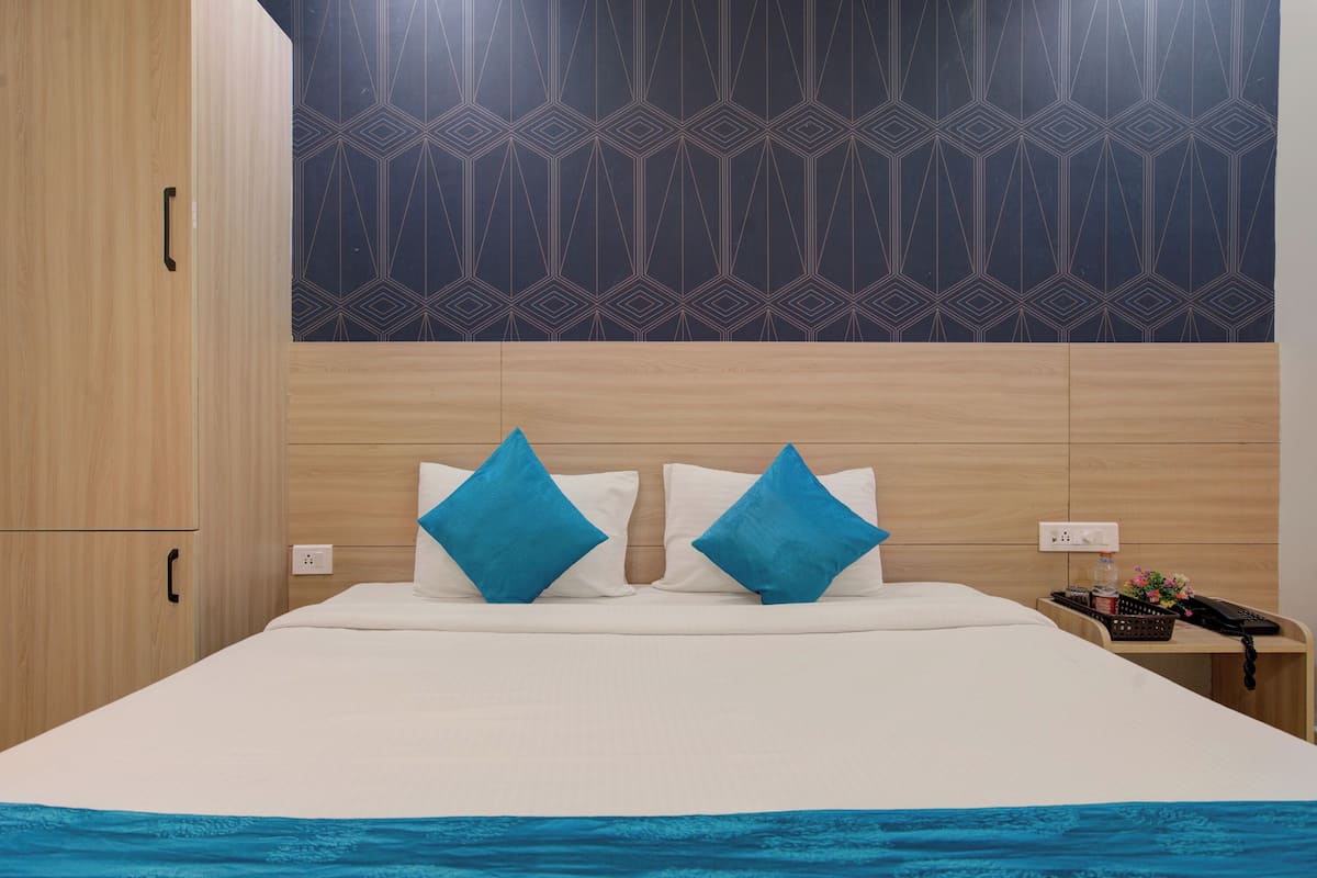 A hotel room with a wooden headboard, a bed with white bedding and two blue pillows, a nightstand with a phone and a small plant.