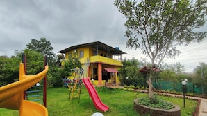A playground with a yellow slide and a red slide, a tree, and a building with a red roof in the background. - My Village Home (Murbad)