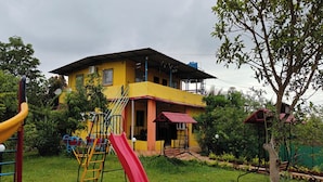 A yellow building with a red slide, surrounded by greenery and a playground. - My Village Home (Murbad)
