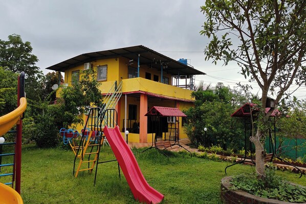 A yellow building with a red slide, surrounded by greenery and a playground. - My Village Home (Murbad)