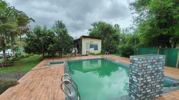 A swimming pool with a wooden deck, a small building with a sign, and surrounding greenery. - My Village Home (Murbad)