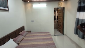 A bedroom with a bed, a clock, and a door leading to another room. - My Village Home (Murbad)