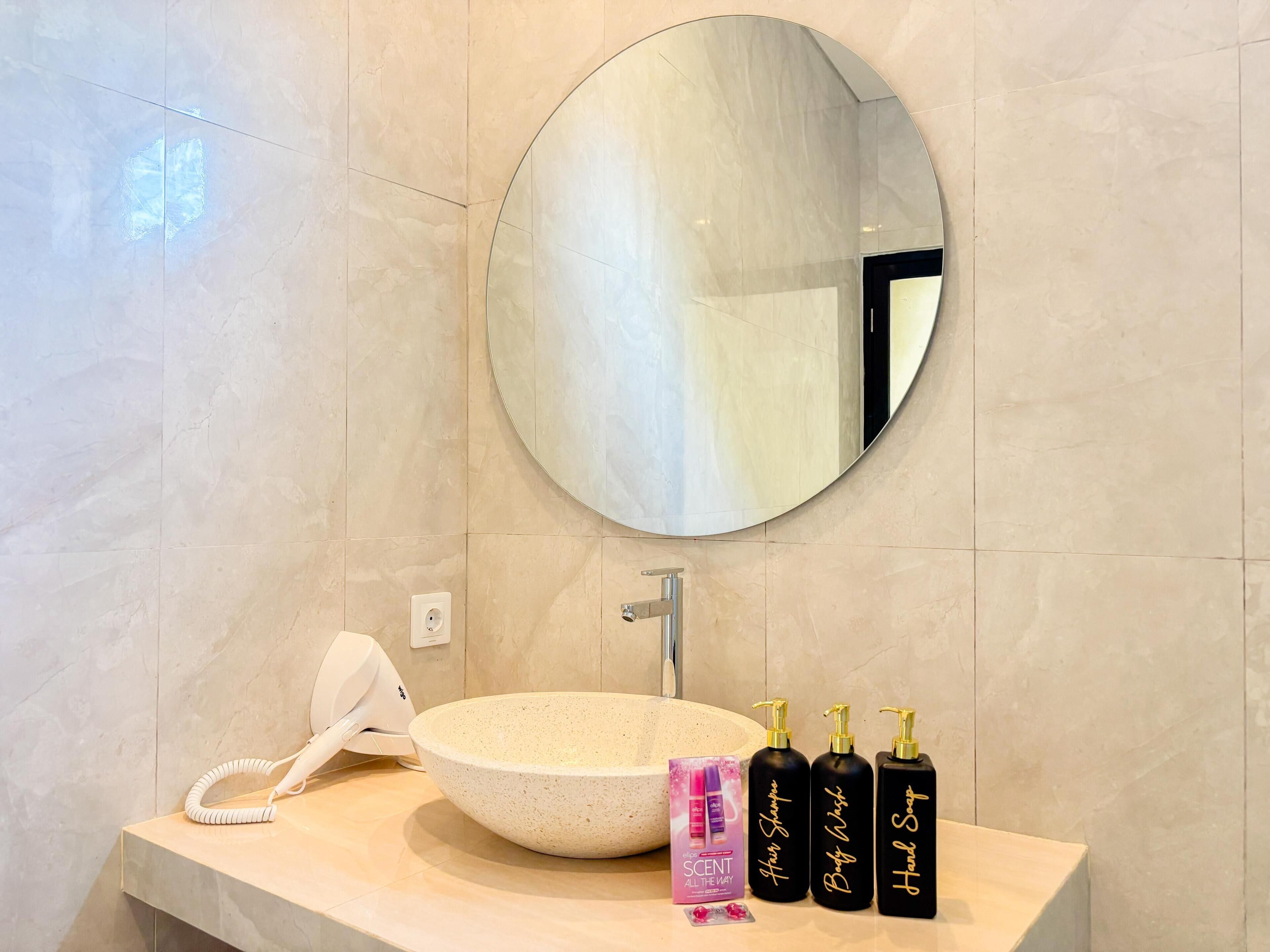 Un bagno con uno specchio rotondo, un lavandino bianco e tre flaconi neri etichettati 'Hair Shampoo', 'Body Wash' e 'Hand Soap'.