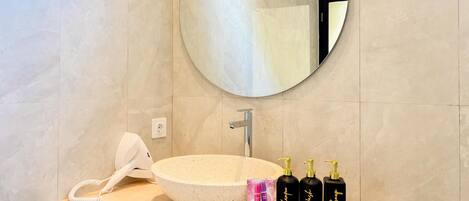 Un bagno con uno specchio rotondo, un lavandino bianco e tre flaconi neri etichettati 'Hair Shampoo', 'Body Wash' e 'Hand Soap'.