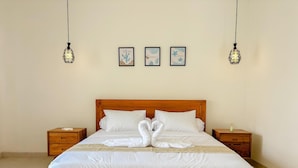 A neatly made bed with white linens and a swan-shaped towel arrangement. There are two bedside tables with lamps, and three framed artworks on the wall above the bed. - Villa Andra Canggu (Canggu)