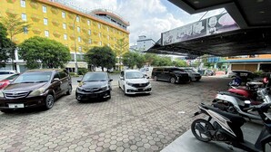 A parking area with several cars and a motorcycle, a yellow building with a geometric pattern, and a billboard with advertisements. - Lucky Star Hotel Near Harbour Bay Batam (Batam)
