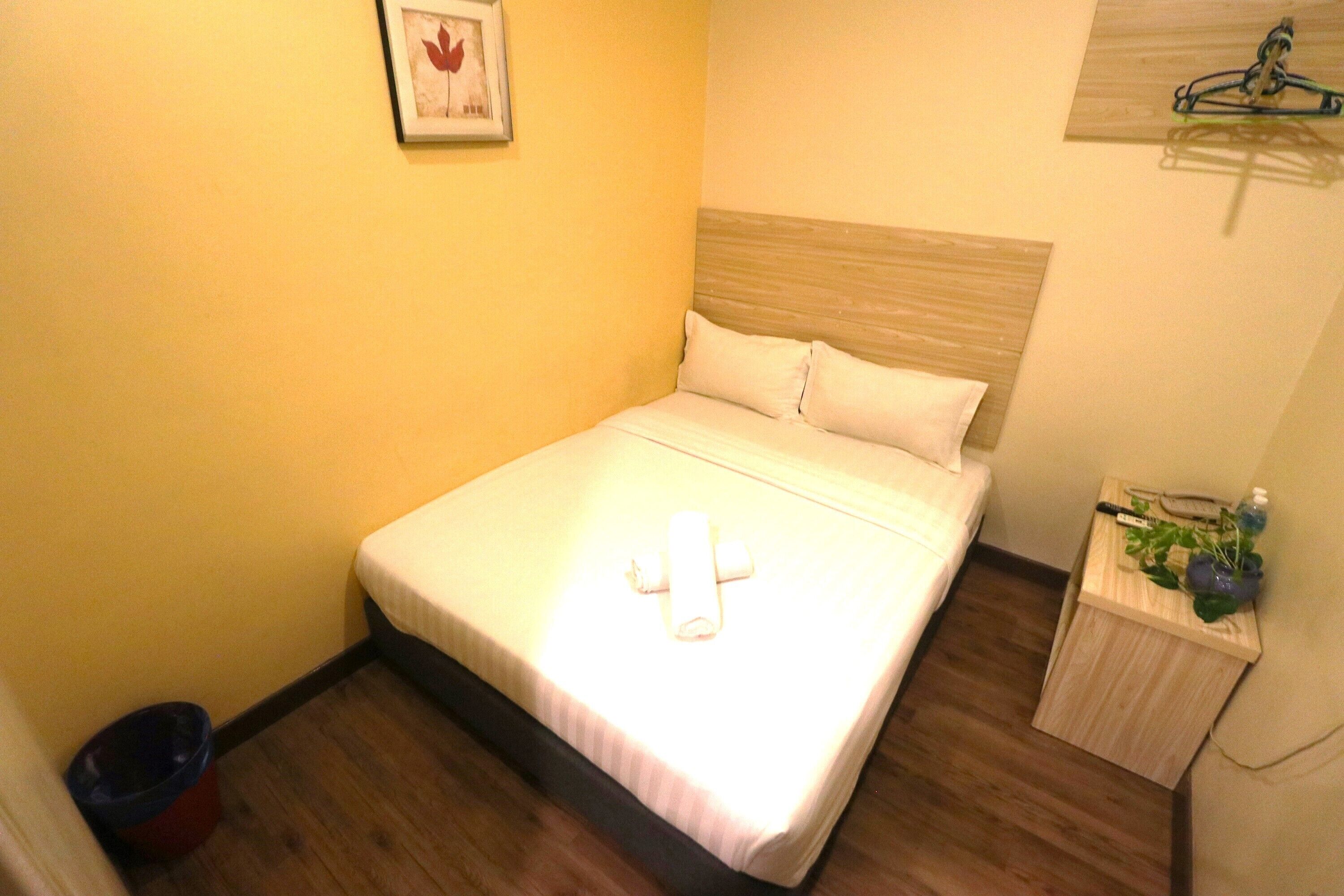 A small, neatly made bed with a white mattress and pillows, a wooden headboard, a small bedside table with a plant, and a framed picture on the wall.
