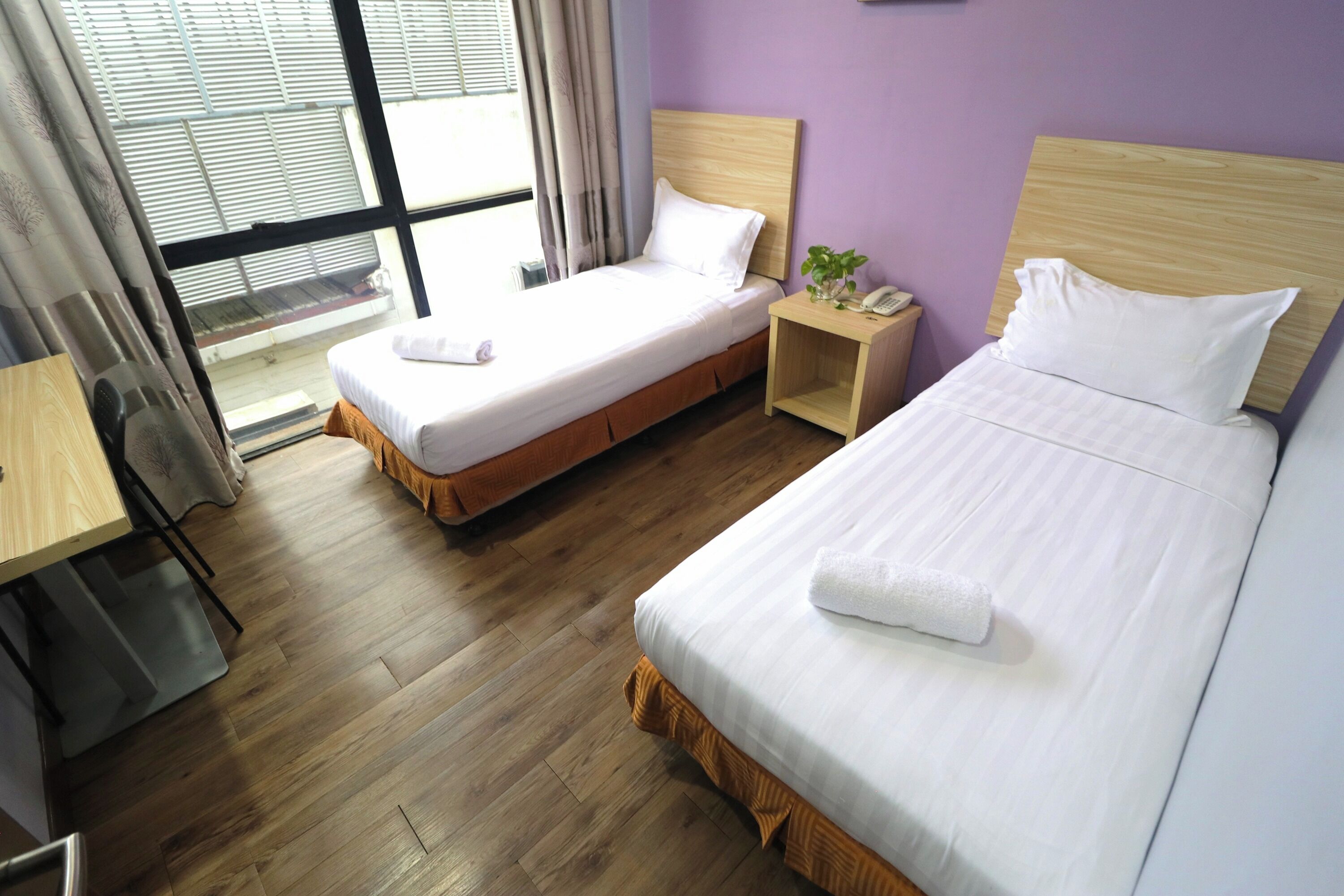 A hotel room with two beds, a wooden floor, a desk, and a window with curtains.