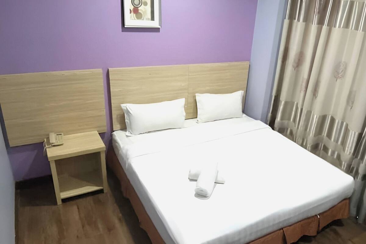 Hotellihuone, jossa on puinen sängynpääty, yksihenkinen sänky valkoisilla vuodevaatteilla, yöpöytä, jossa on puhelin, ja ikkuna, jossa on verhot.