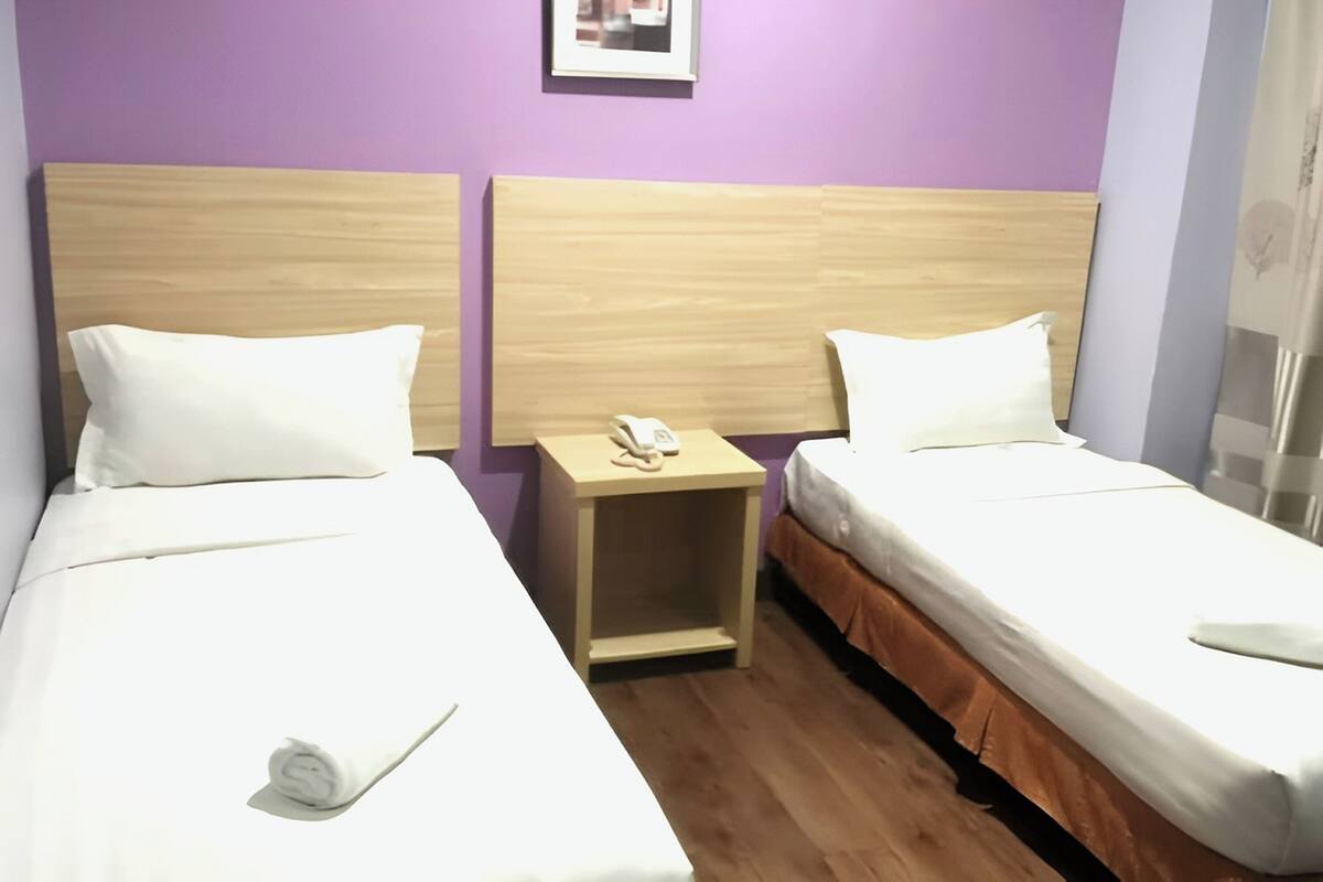 Hotellihuone, jossa on kaksi erillistä sänkyä, puiset päätyosat, valkoiset vuodevaatteet, pieni yöpöytä, jossa on puhelin, ja violetti seinä.