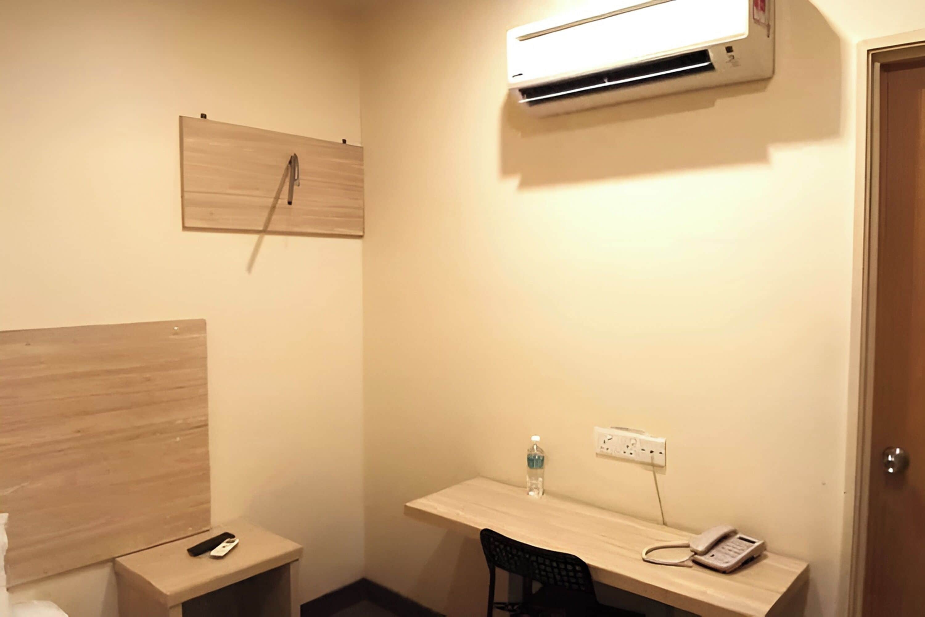A small, well-lit room with a wooden desk, a chair, a bedside table with a remote control, a water bottle, and a phone. An air conditioner is mounted on the wall above the desk.