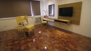 A modern living room with a wooden parquet floor, a yellow armchair, a small round table with a book and a plant, a wooden stool, a flat-screen TV mounted on a wooden panel, and a window with curtains. - Bryan Condo Suites Makati near Airport (Makati)