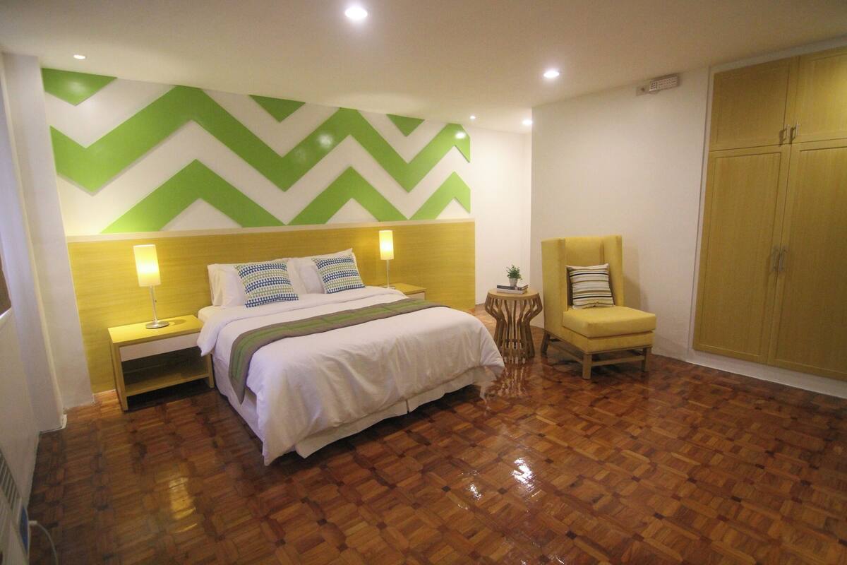 Une chambre à coucher comprenant un lit, des tables de chevet, une chaise, une armoire et un mur décoré d’un motif à chevrons verts et blancs.