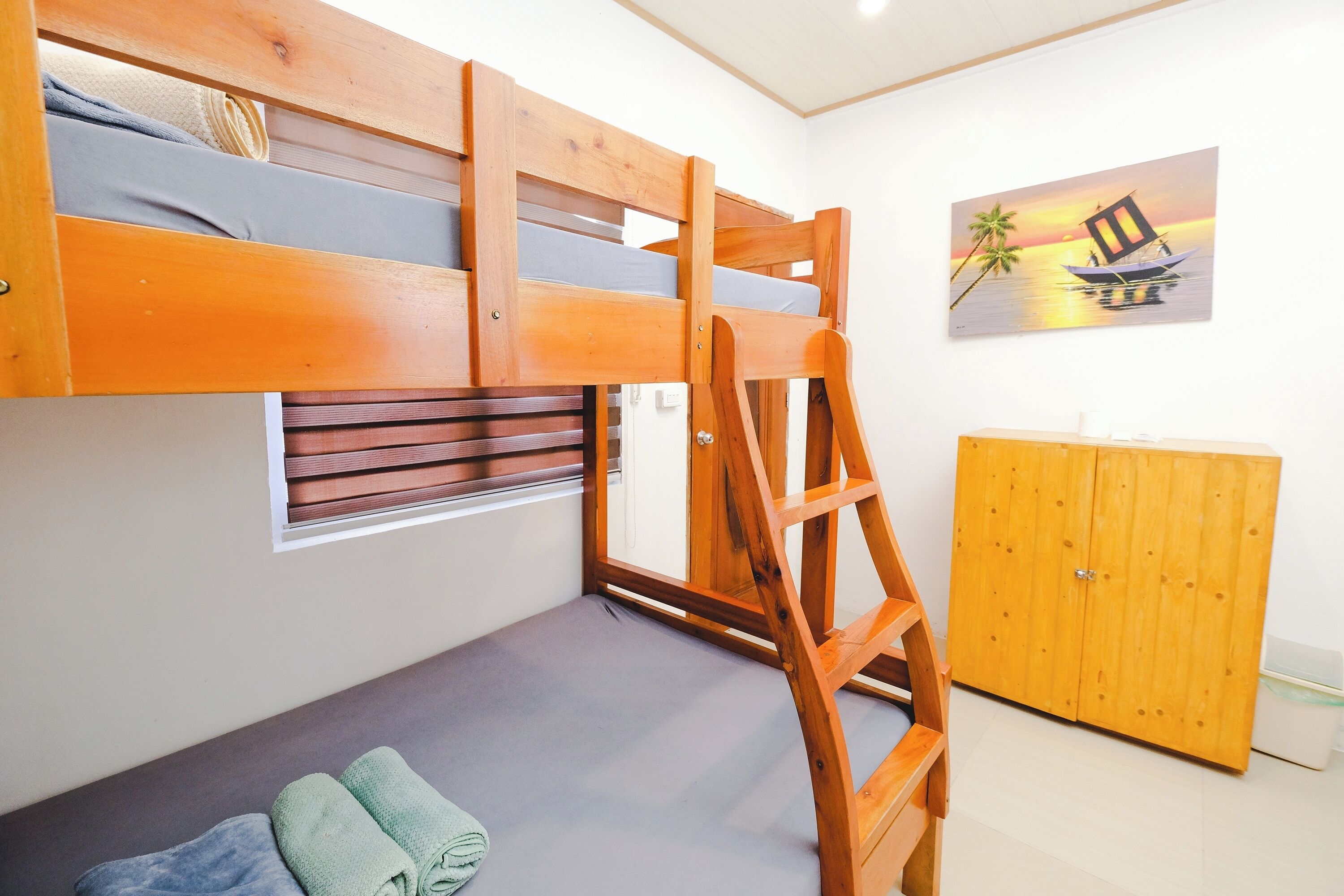 Un lit superposé avec une échelle, une petite fenêtre, une armoire en bois et un tableau représentant un bateau sur l’eau.