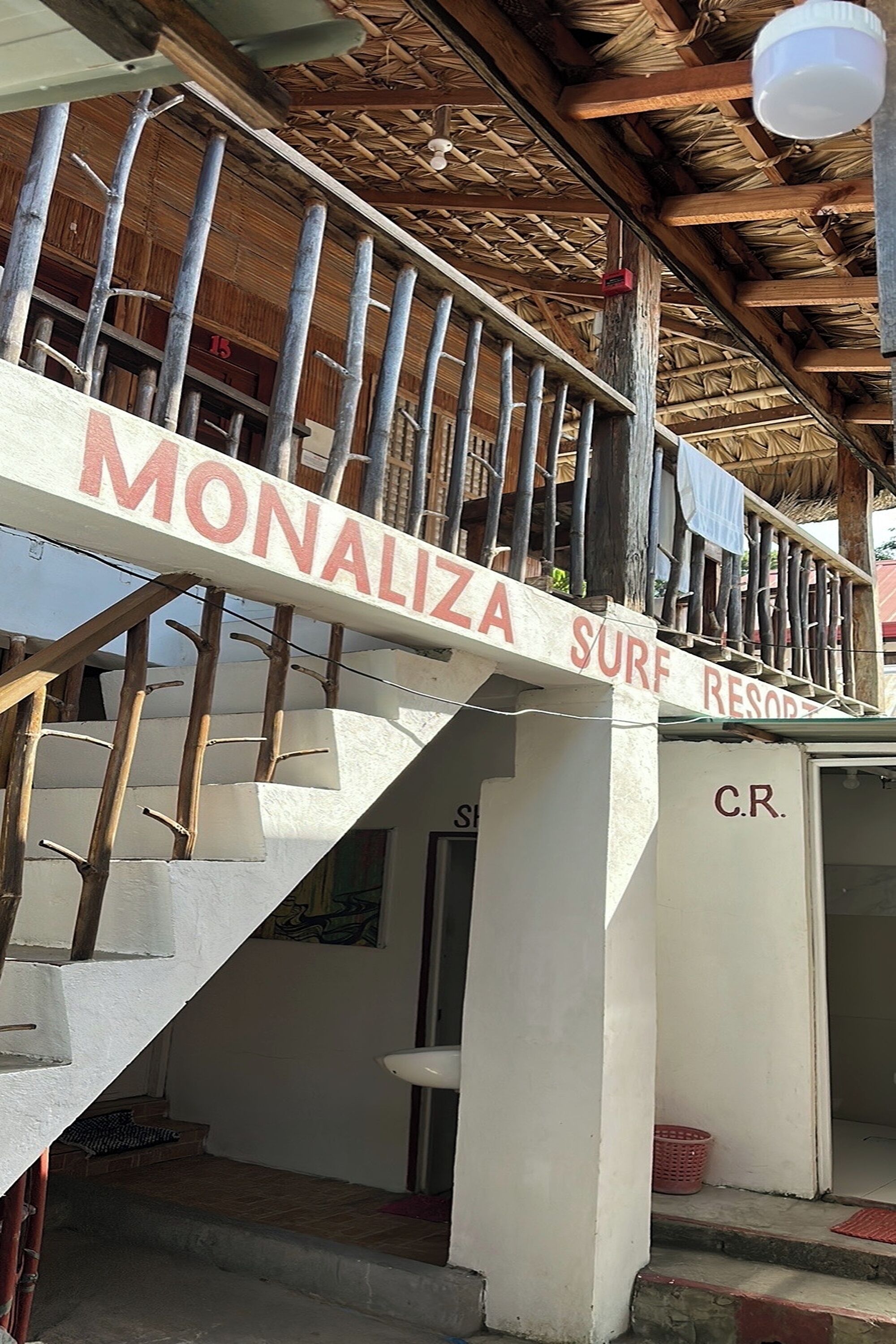 Eine Treppe führt zu einem Gebäude mit dem Schriftzug „Monaliza Surf Resort“, dessen Dach mit Schilf gedeckt und die Geländer aus Bambus sind.