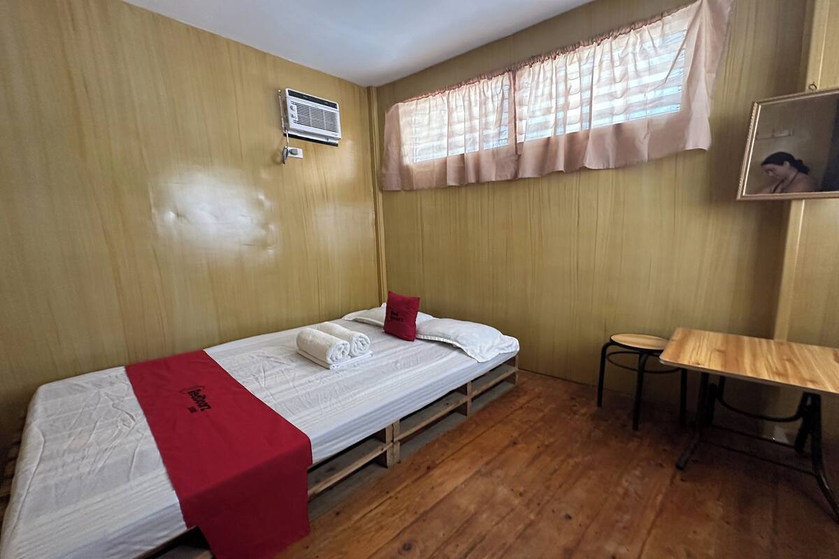 A single bed with a red blanket, a small table, and a window with curtains.