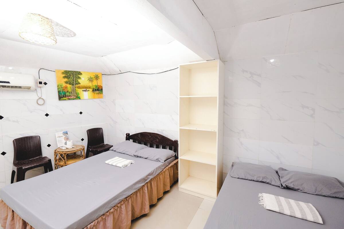 A hotel room with a bed, a small table with chairs, a bookshelf, and a painting on the wall.