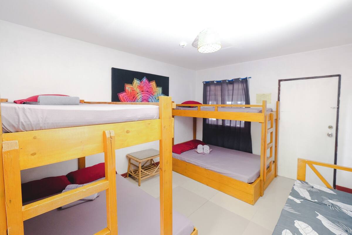A dormitory room with bunk beds, a small wooden stool, a colorful artwork on the wall, and a door with a curtain.