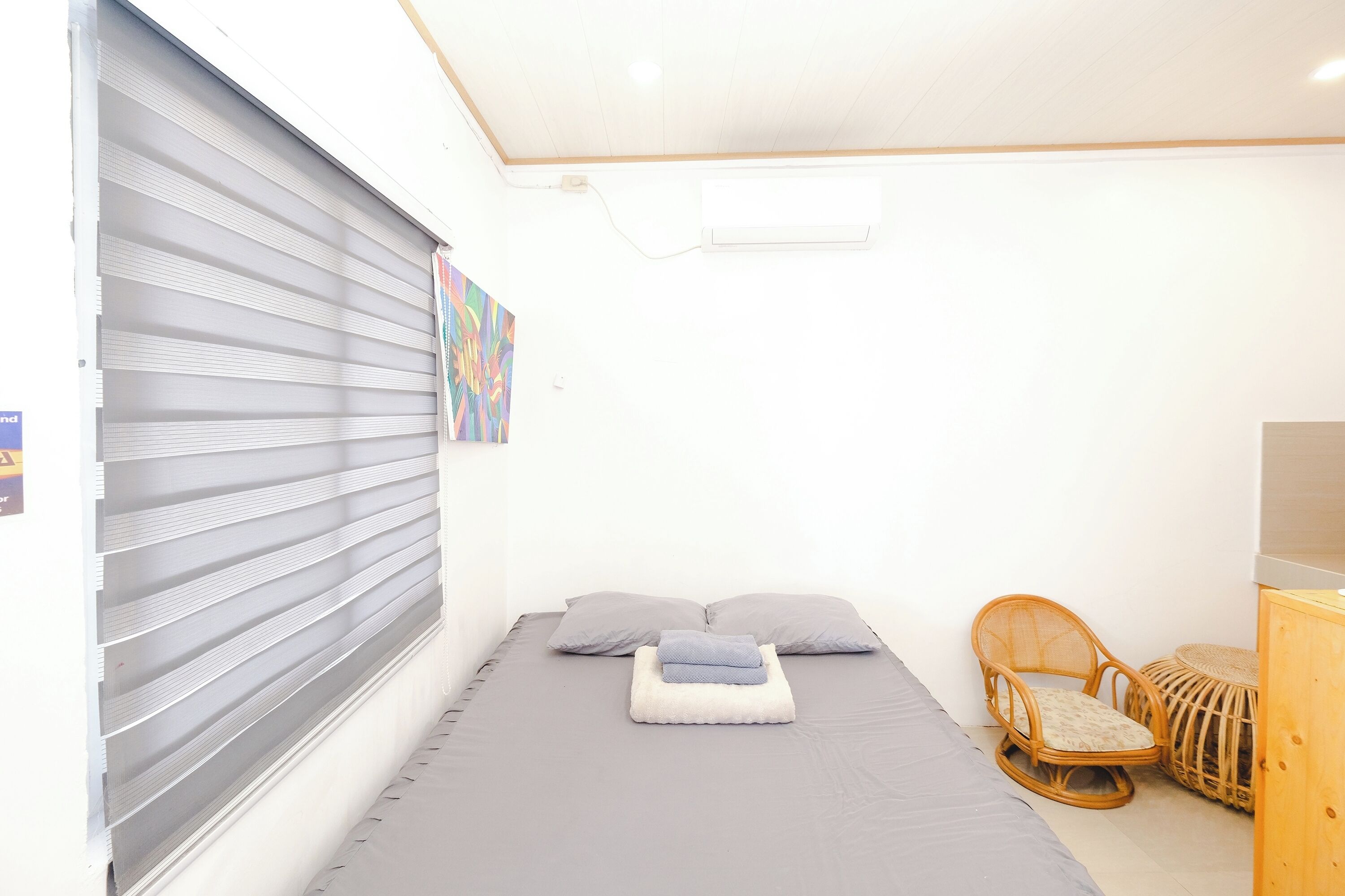 A small, neatly arranged room with a bed, two pillows, a chair, and a small wooden cabinet.