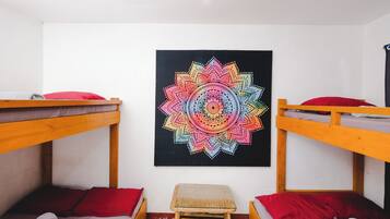 A dormitory room with two bunk beds, a small stool, and a colorful mandala artwork on the wall.