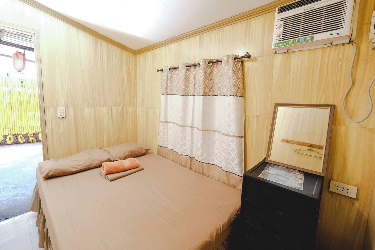 A room with a bed, a window with curtains, a wall-mounted air conditioner, and a small table with a lamp.