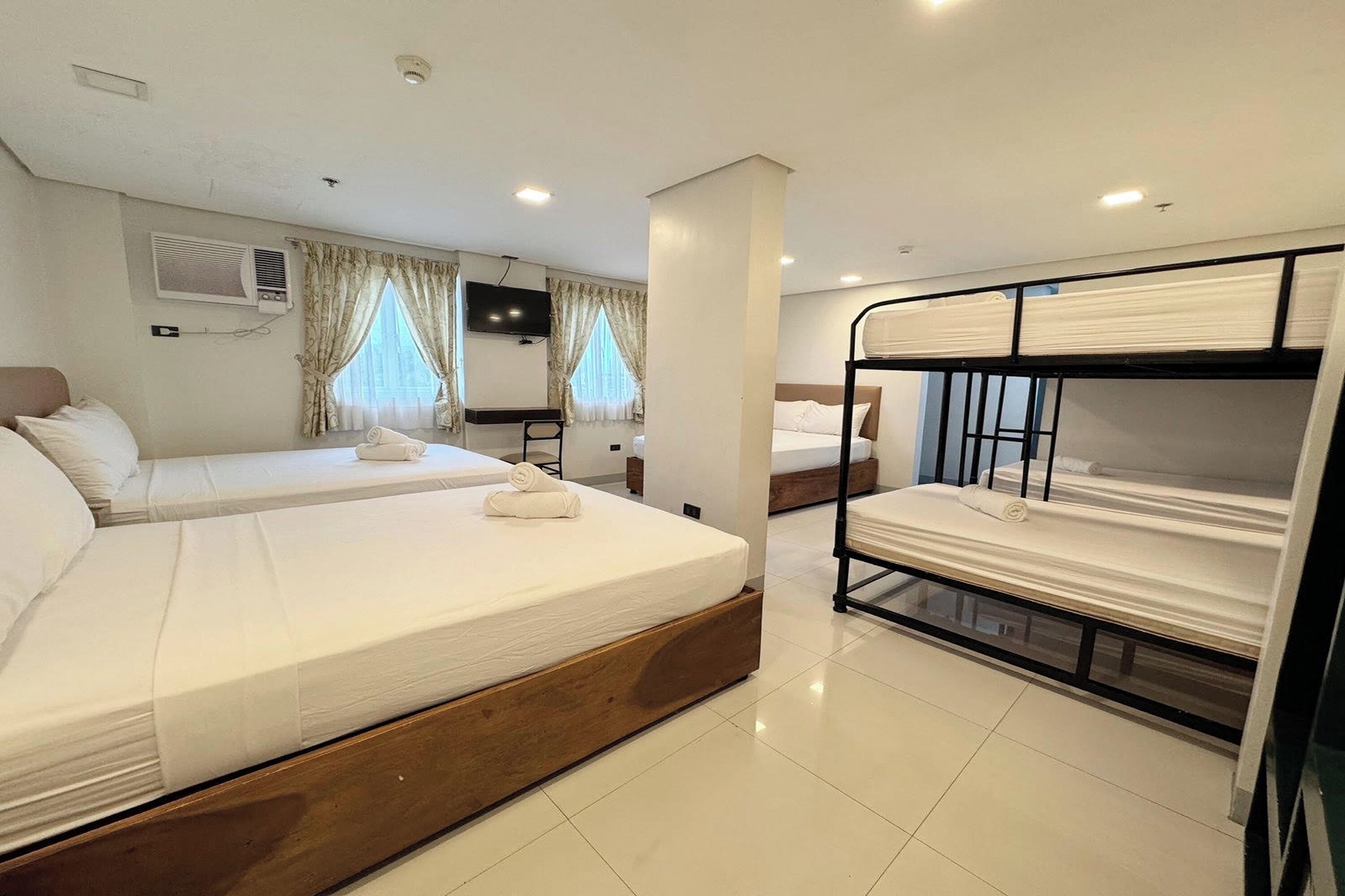 A hotel room with two single beds, a bunk bed, a TV, an air conditioning unit, and curtains on the windows.