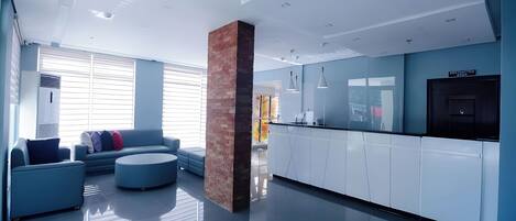 A modern hotel lobby with a blue sofa, a round ottoman, a kitchen area, and a brick pillar.