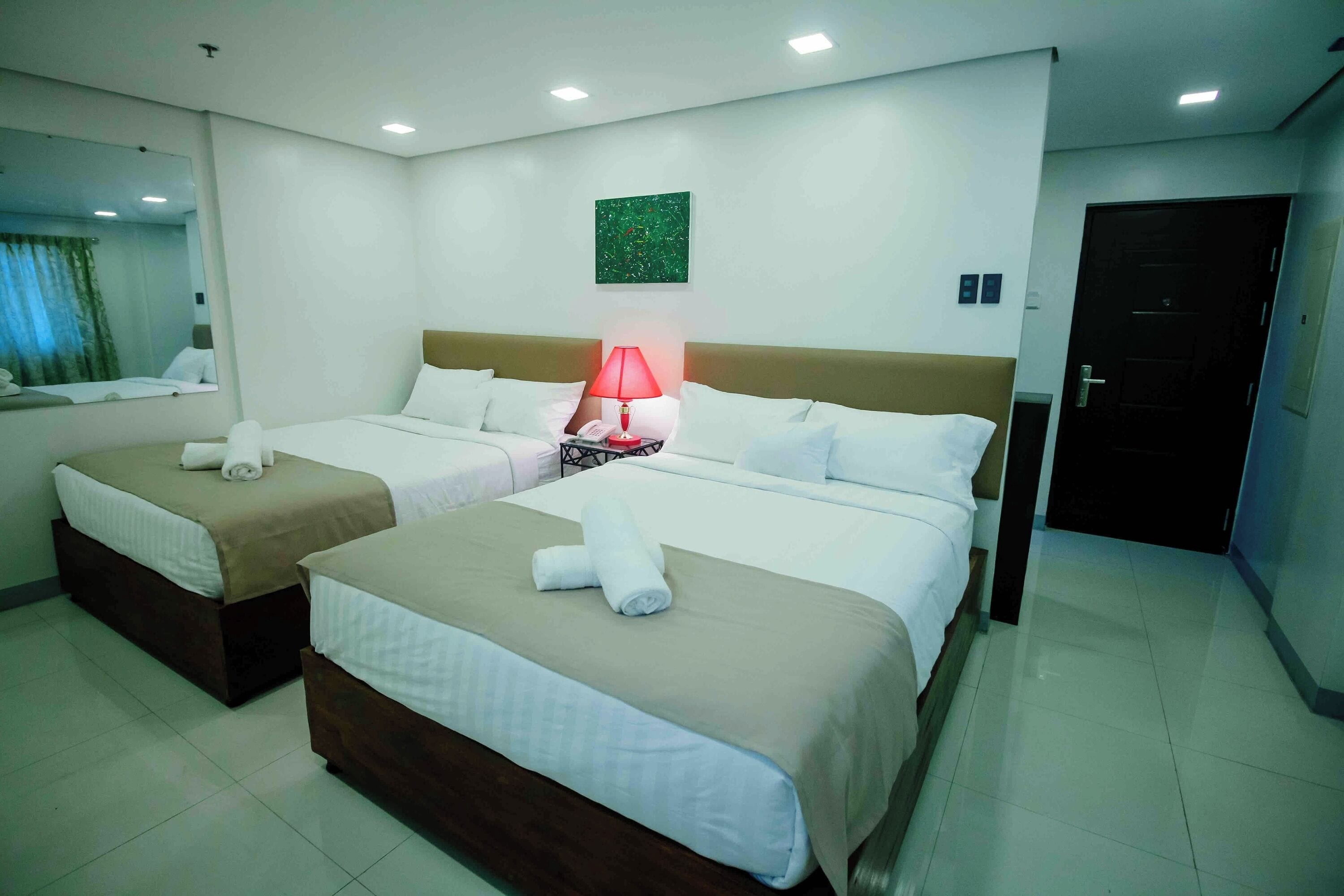Ein Hotelzimmer mit zwei Betten, einem Nachttisch mit Lampe, einem Spiegel und einem grünen, abstrakten Gemälde an der Wand.