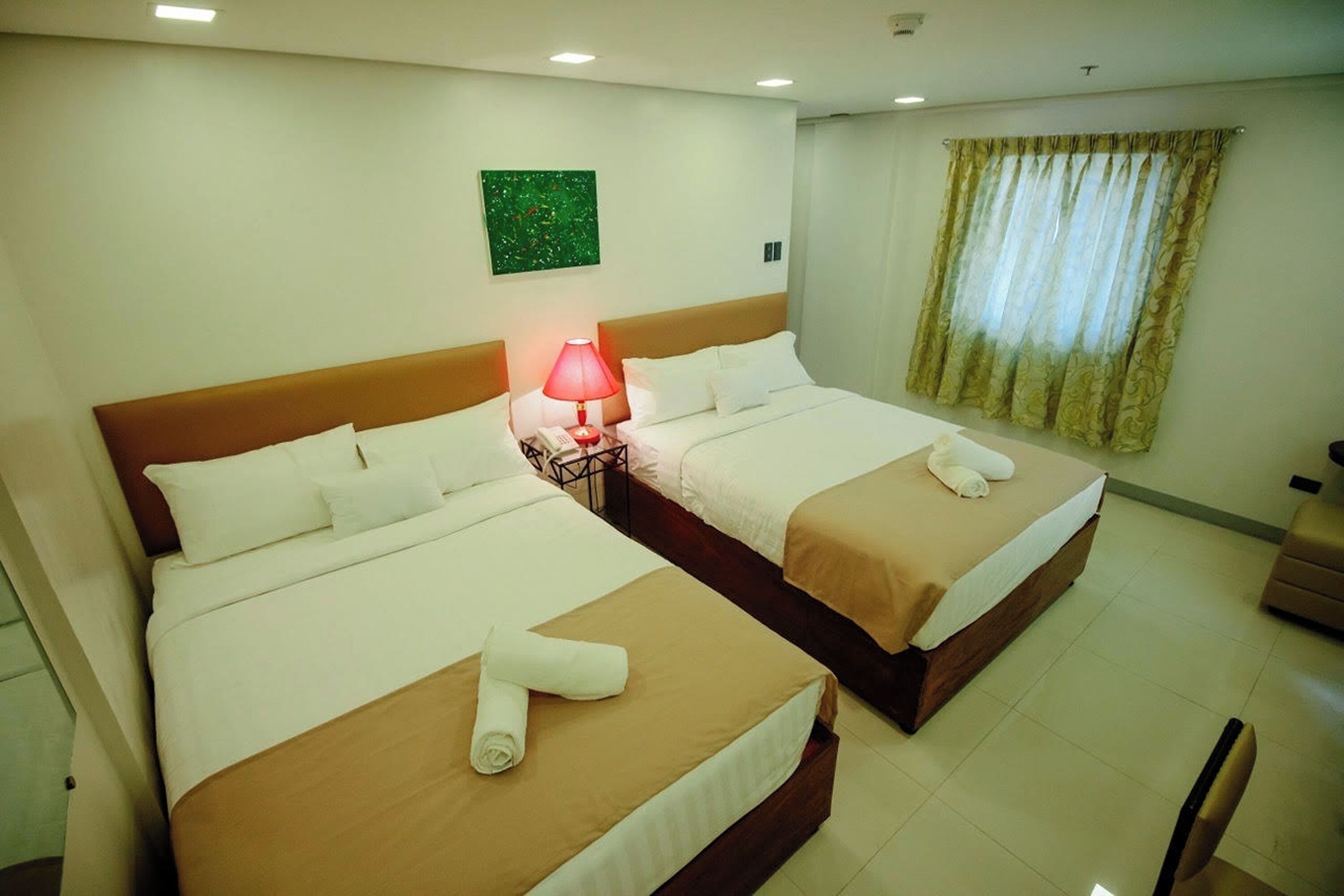 A hotel room with two beds, a nightstand with a lamp, a green abstract painting, and a window with curtains.