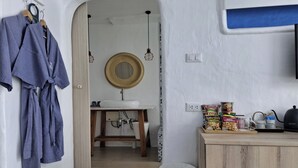 Une pièce avec un meuble en bois, un évier et un miroir au mur. Des vêtements sont suspendus à un portant et une poubelle est posée sur le sol.