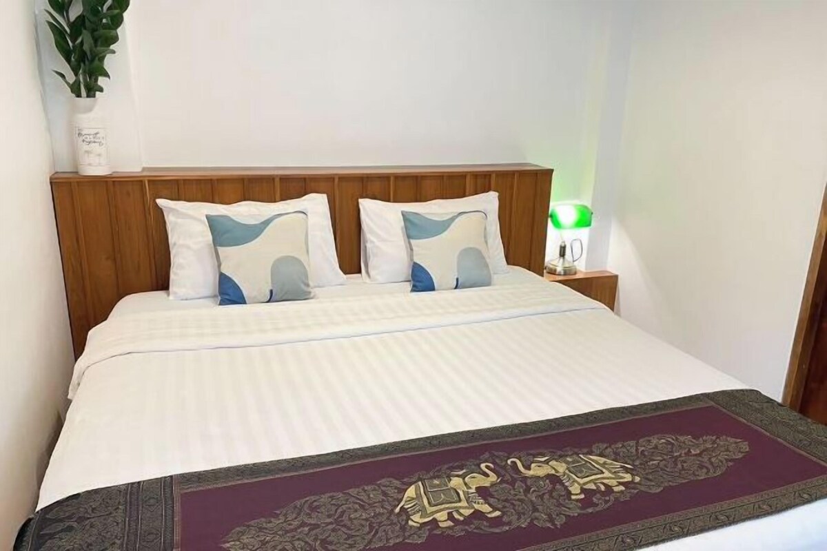 Una cama bien hecha con una colcha blanca y azul estampada, un plaid decorativo al pie de la cama y una mesita de noche con una lámpara verde.