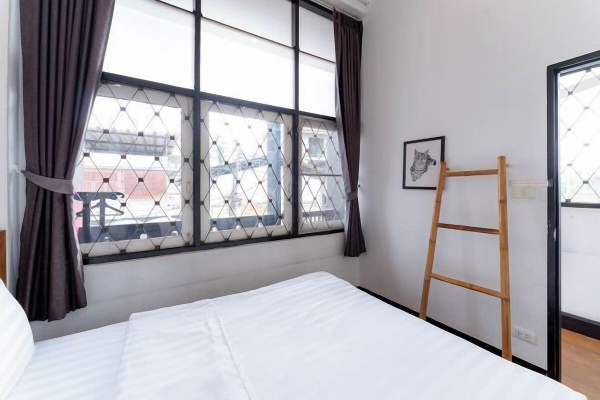A hotel room with a bed, a window with curtains, a ladder, and a framed picture on the wall.