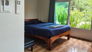 A bedroom with a wooden bed, a bedside table, a nightstand, a window with a view of greenery, and a wall-mounted thermostat. - 99 House (Pattaya)