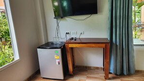 A compact room with a flat-screen TV mounted on the wall, a small refrigerator, a wooden table, and a window with a view of greenery. - 99 House (Pattaya)