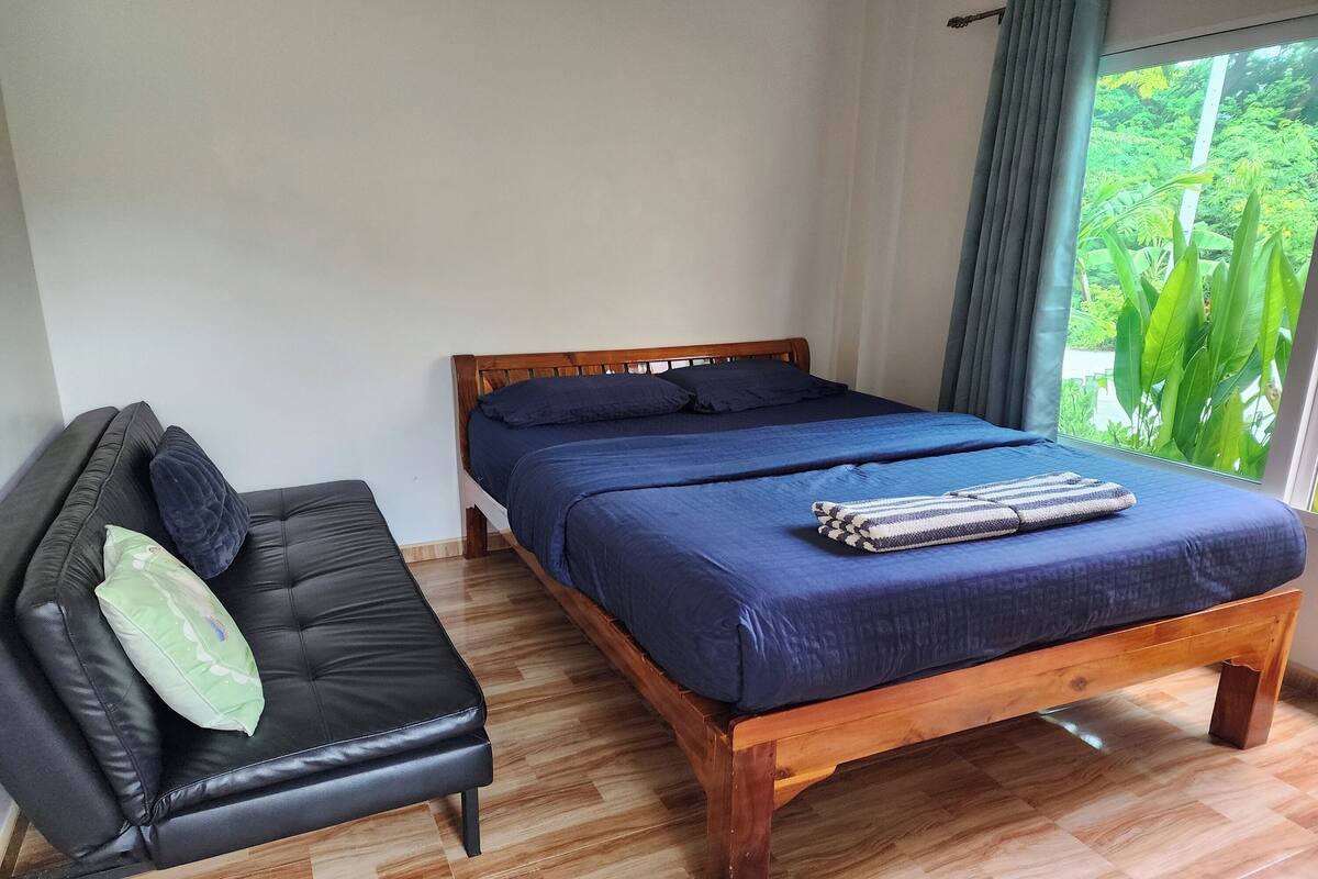Une chambre avec un lit en bois, un canapé en cuir noir et une grande fenêtre donnant sur un paysage verdoyant.