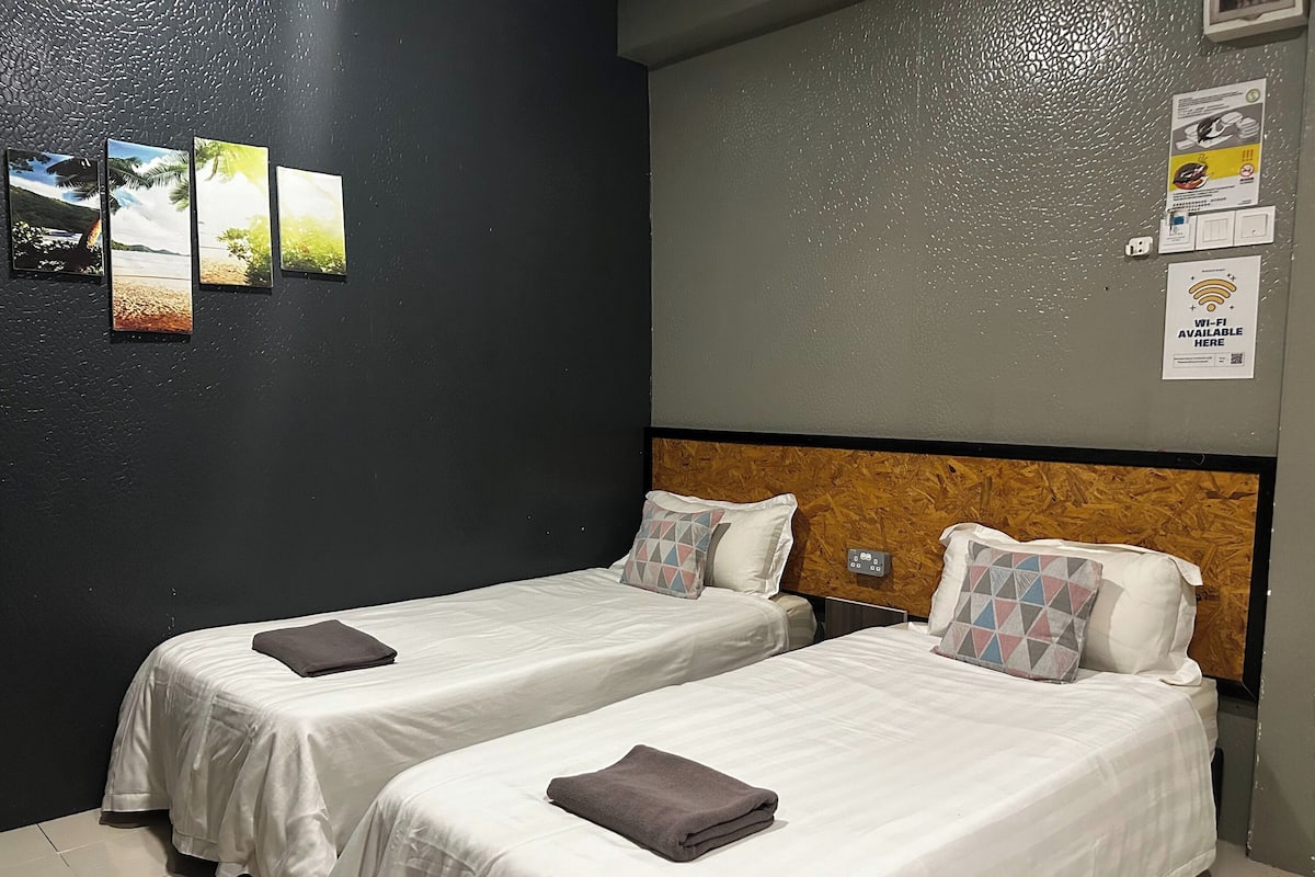 A hotel room with two single beds, a wooden headboard, a wall-mounted power outlet, and a Wi-Fi sign.