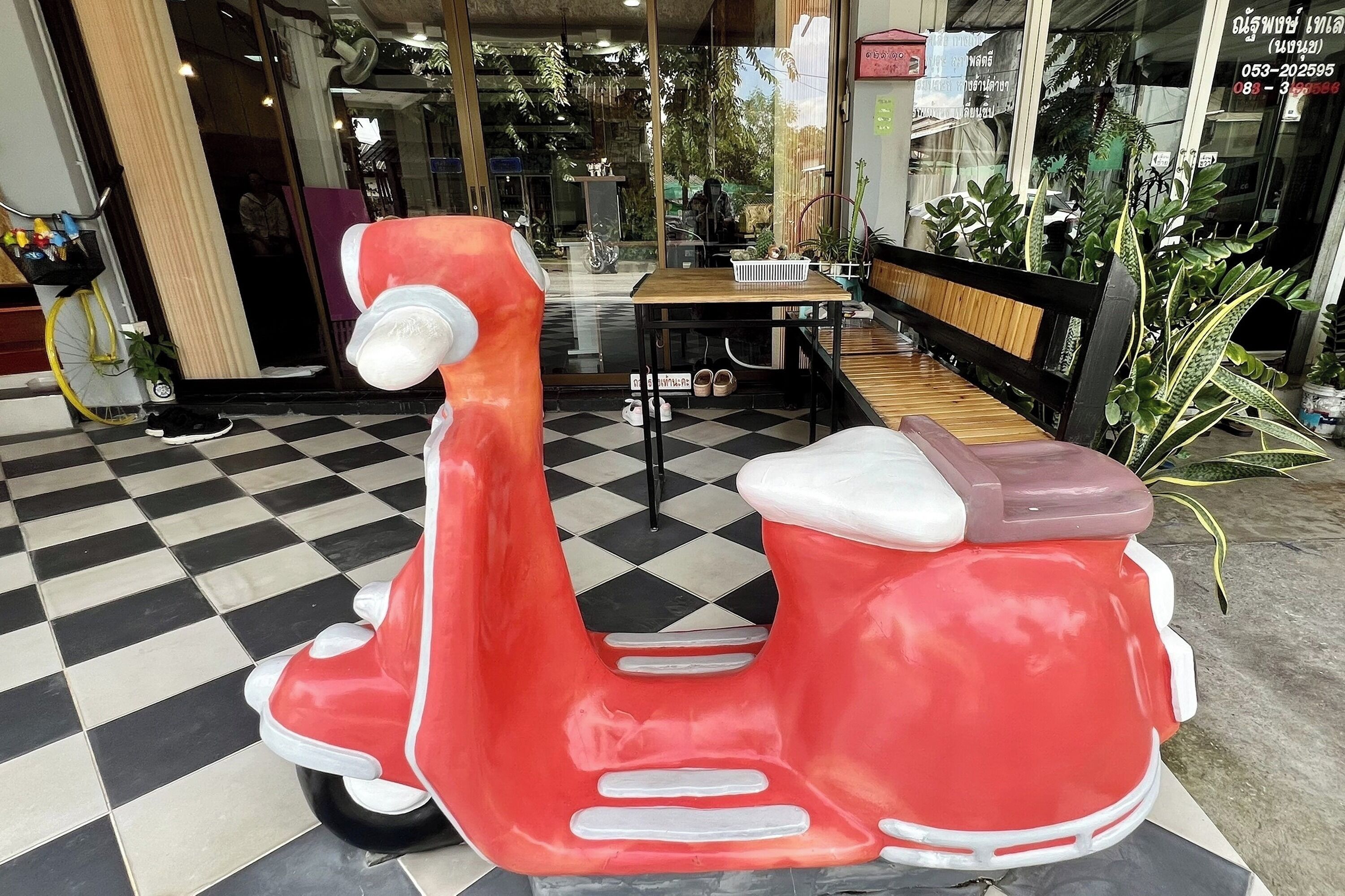 Una patineta roja estacionada frente a un edificio con piso de tablones, un banco de madera y plantas en macetas.
