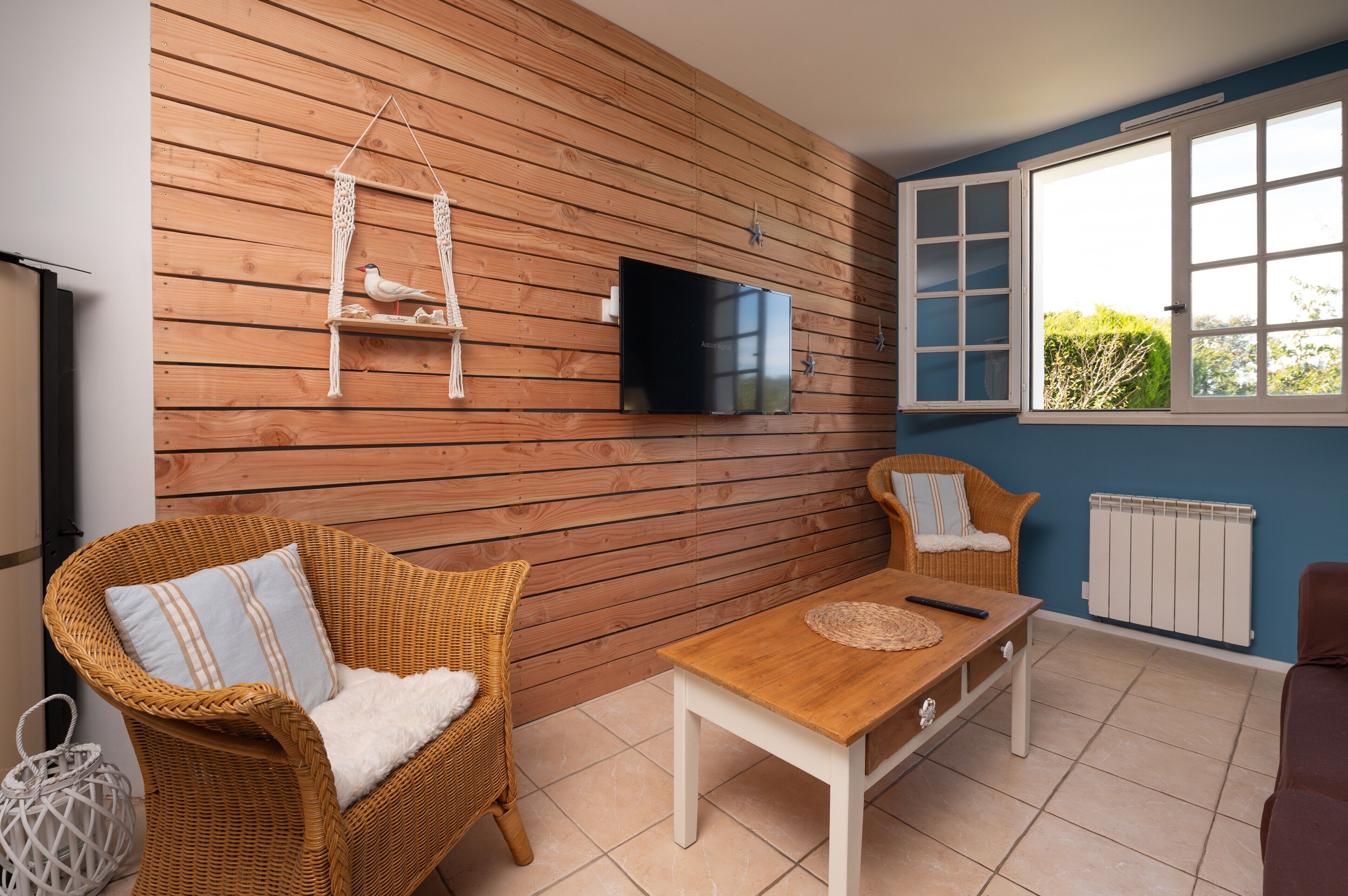 A room with a wooden wall, a television, wicker furniture, a window with a view, and a radiator.