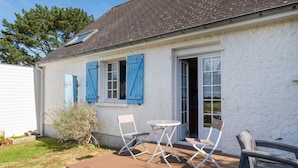 Een wit gebouw met blauwe luiken, een terras met een tafel en stoelen, en een deur die leidt naar een overdekte ingang.