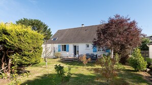 Een huis met een witte gevel, blauwe luiken, een schuin dak en een kleine voortuin met zitplaatsen.