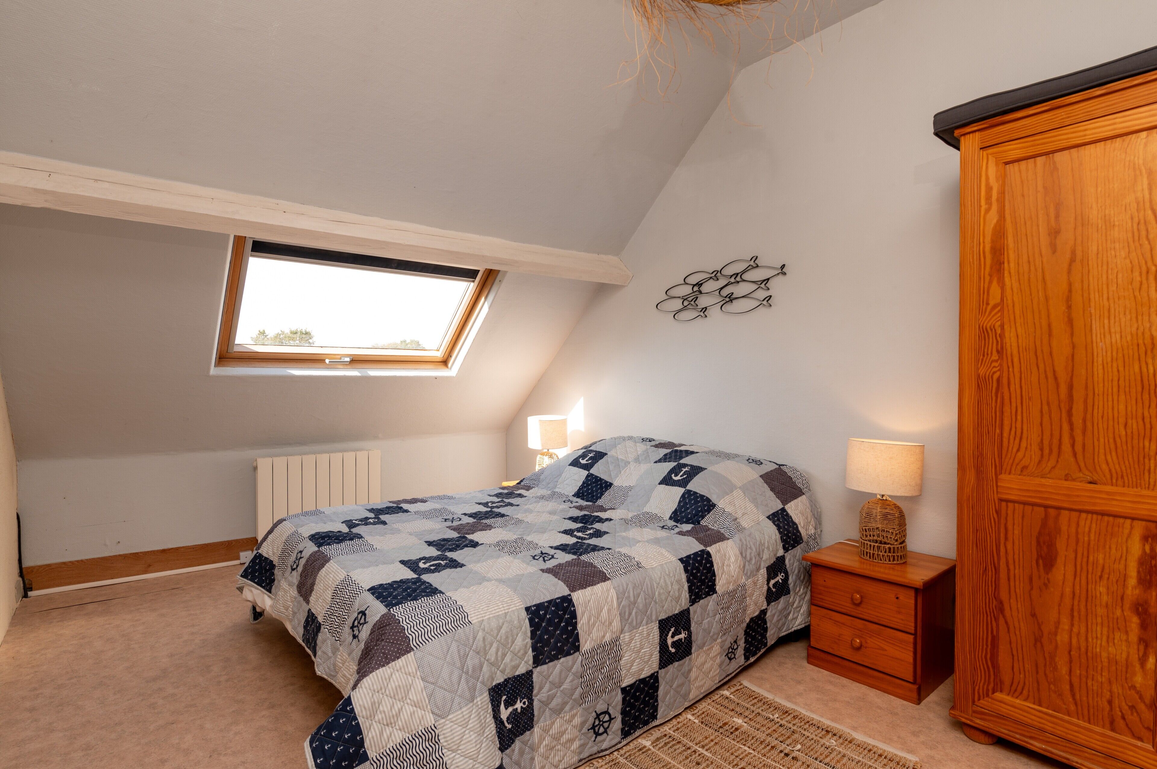 A bedroom with a sloped ceiling, a skylight, a bed with a nautical-themed quilt, a nightstand with a lamp, and a wooden wardrobe.