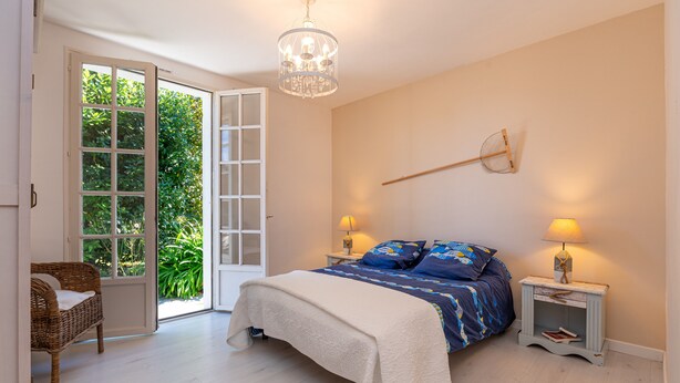 Een slaapkamer met een bed, een rieten stoel, een kroonluchter en uitzicht op groen door een deur.