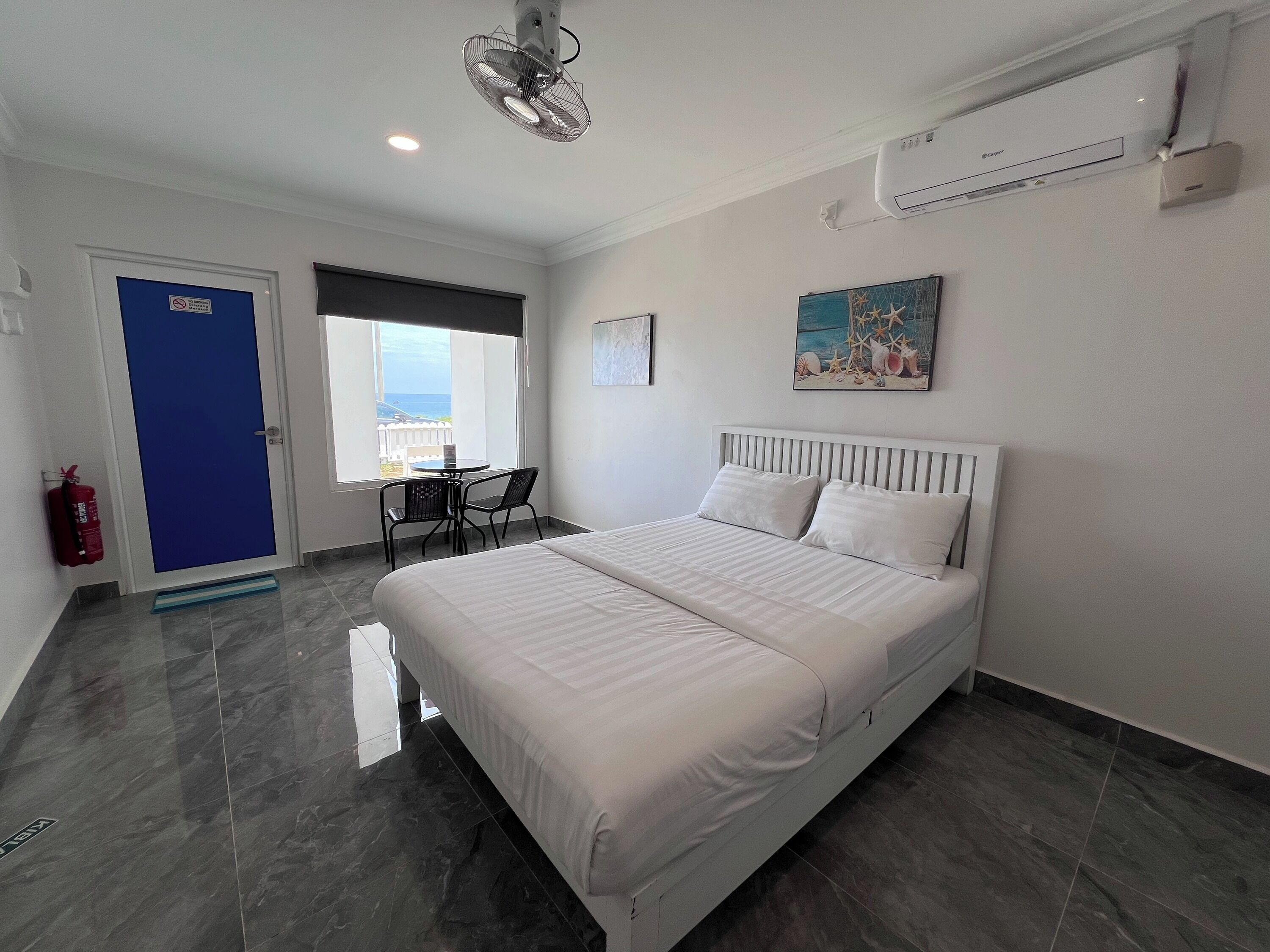 A hotel room with a bed, a small table with two chairs, a window with a view of the sea, a ceiling fan, and an air conditioning unit.