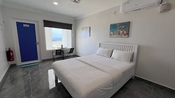 A hotel room with a bed, a small table with two chairs, a window with a view of the sea, a ceiling fan, and an air conditioning unit.