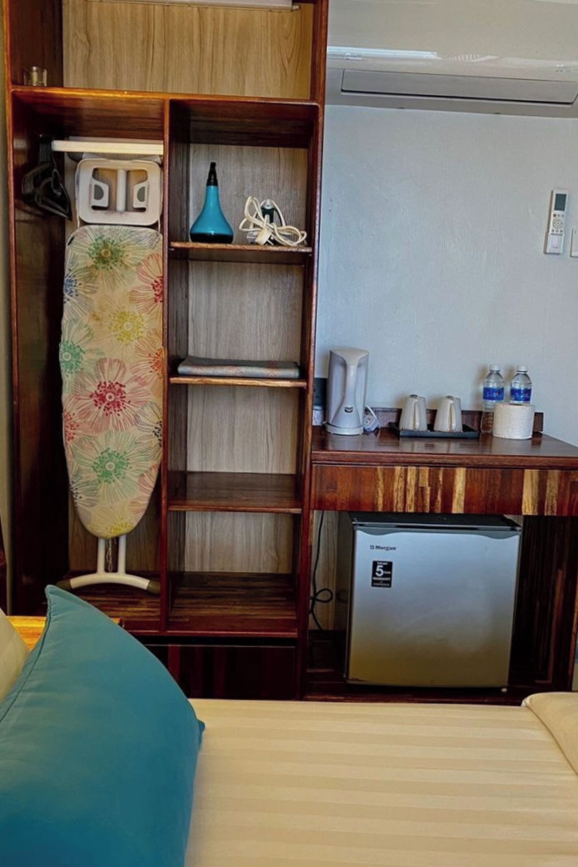 Habitación compacta con cama, kitchenette pequeña, refrigerador, estante con lámpara azul y un depurador de prendas colgante.