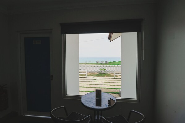 Habitación con vista a una playa y una valla blanca. Hay una puerta con un letrero, una mesa redonda con una silla y una pequeña planta.